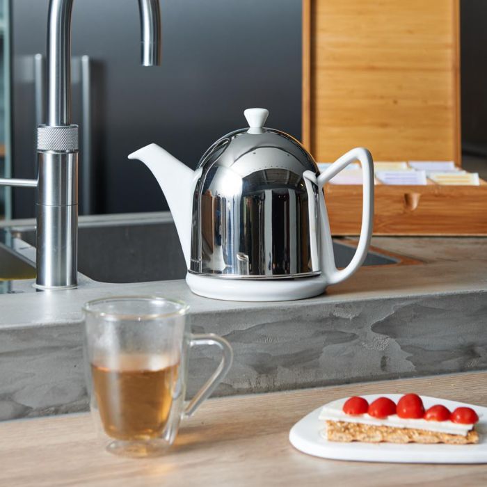 White teapot with a stainless steel cosy on a kitchen counter beside a cup of tea