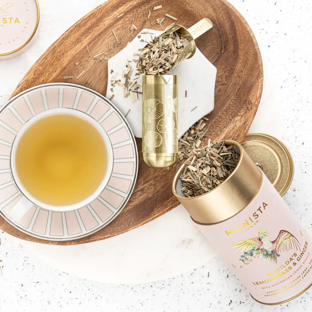 Gold spoon scooping dried chamomile flowers beside rows of Monista tea canisters