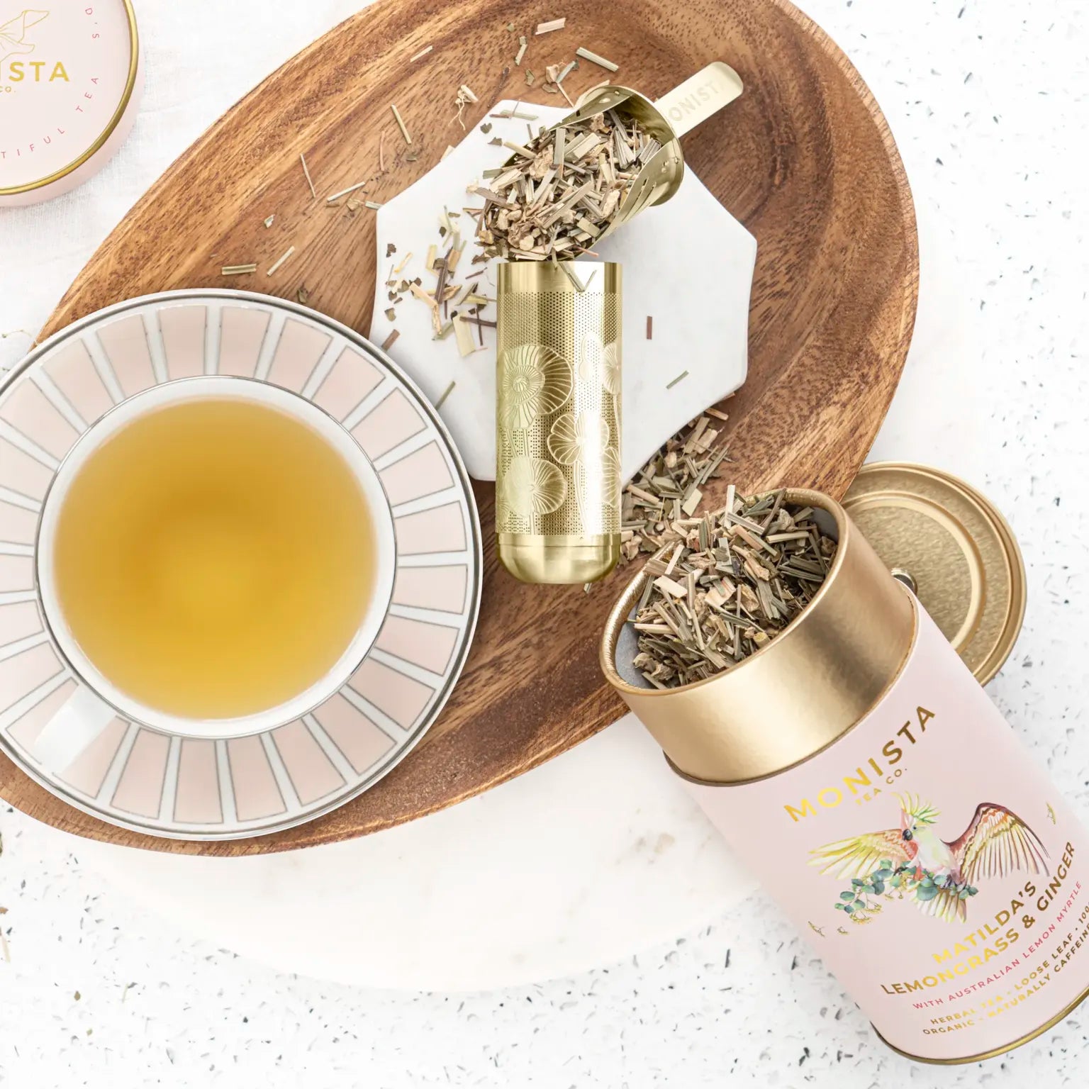 Gold spoon scooping dried chamomile flowers beside rows of Monista tea canisters