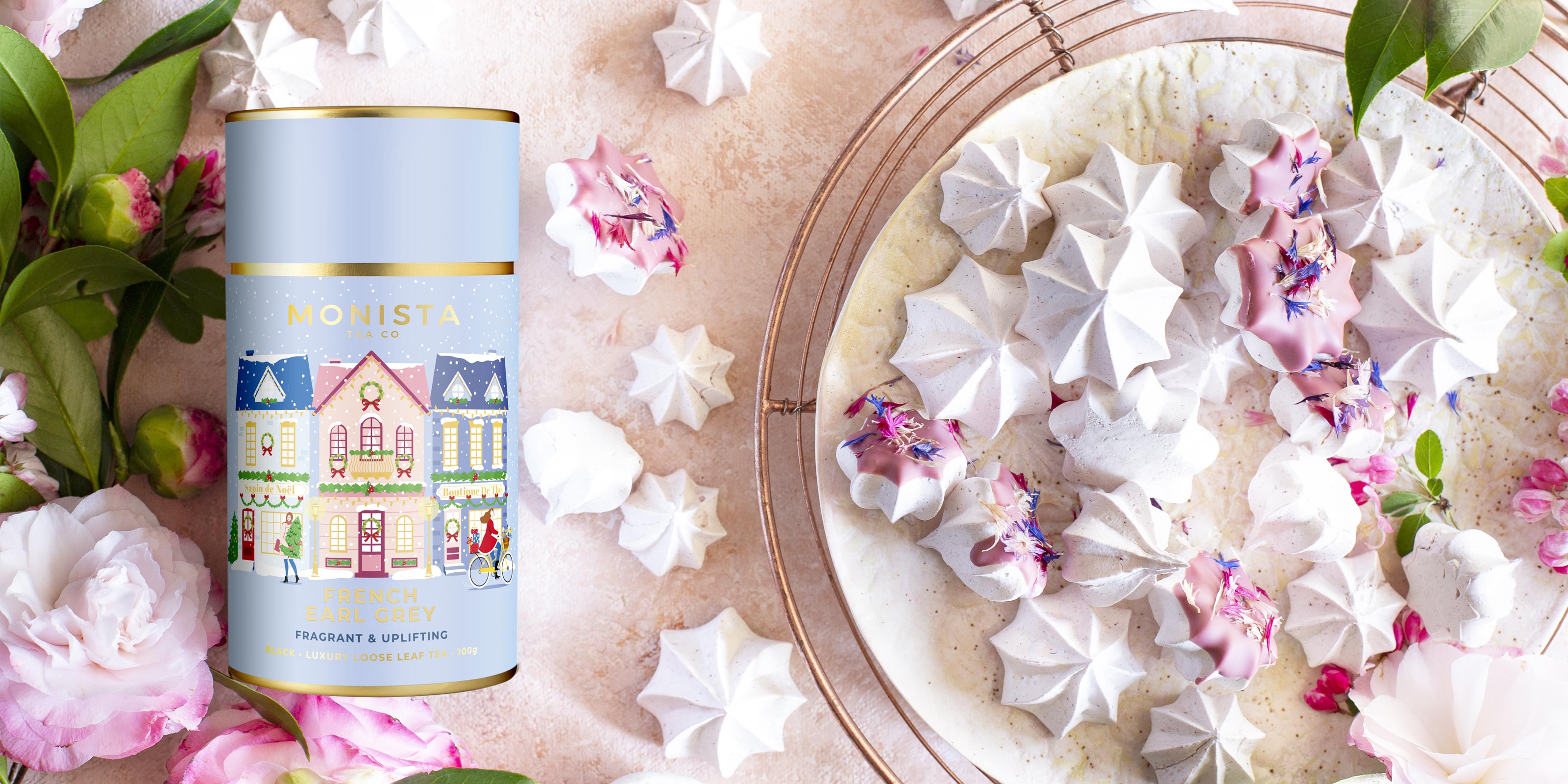 A cylindrical tin of Monista Tea sits beside a plate of white meringue cookies decorated with edible flowers on a pink surface with scattered meringues and green leaves.
