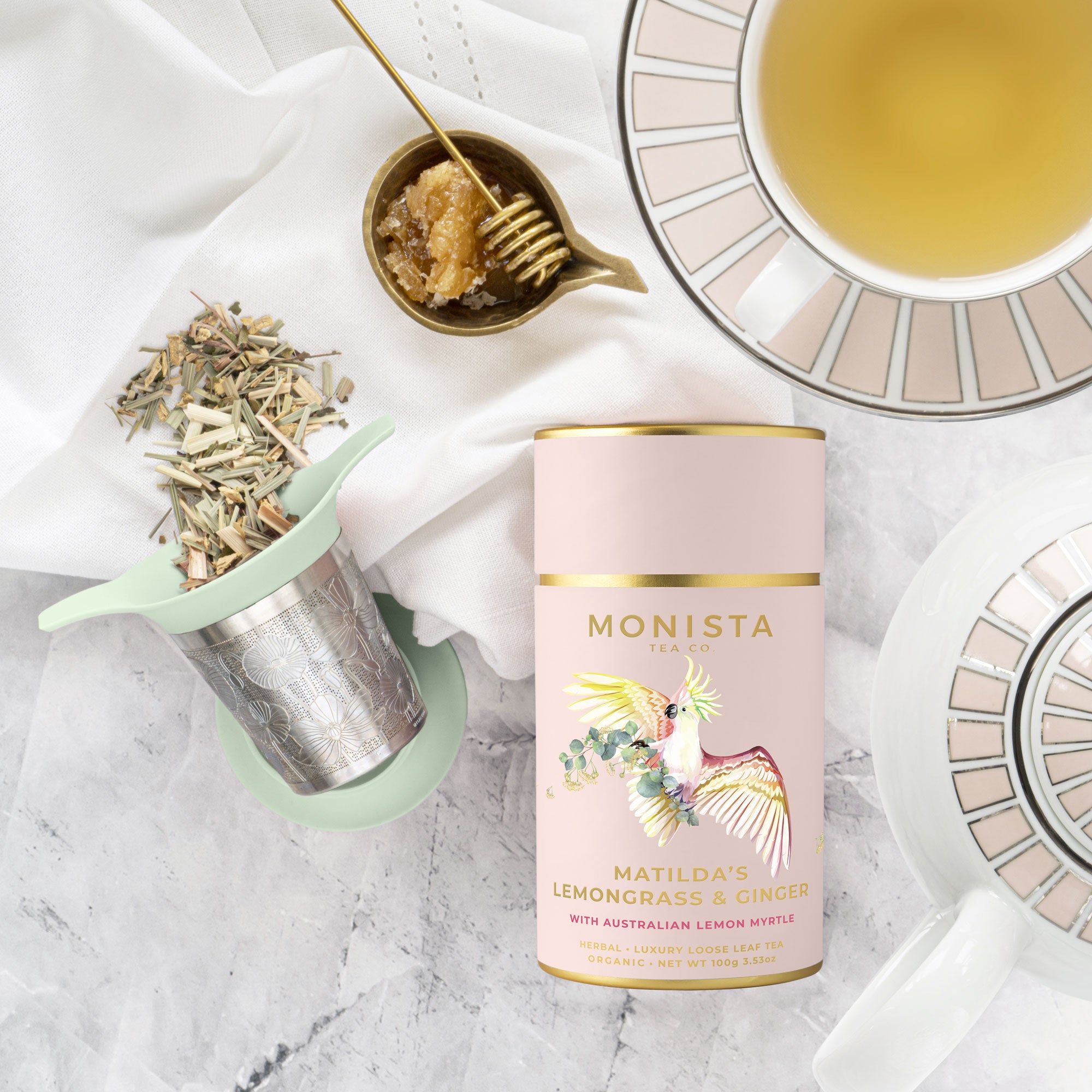 Mint green tea infuser with lemongrass and ginger tea, honey, and a cup of tea on a marble surface