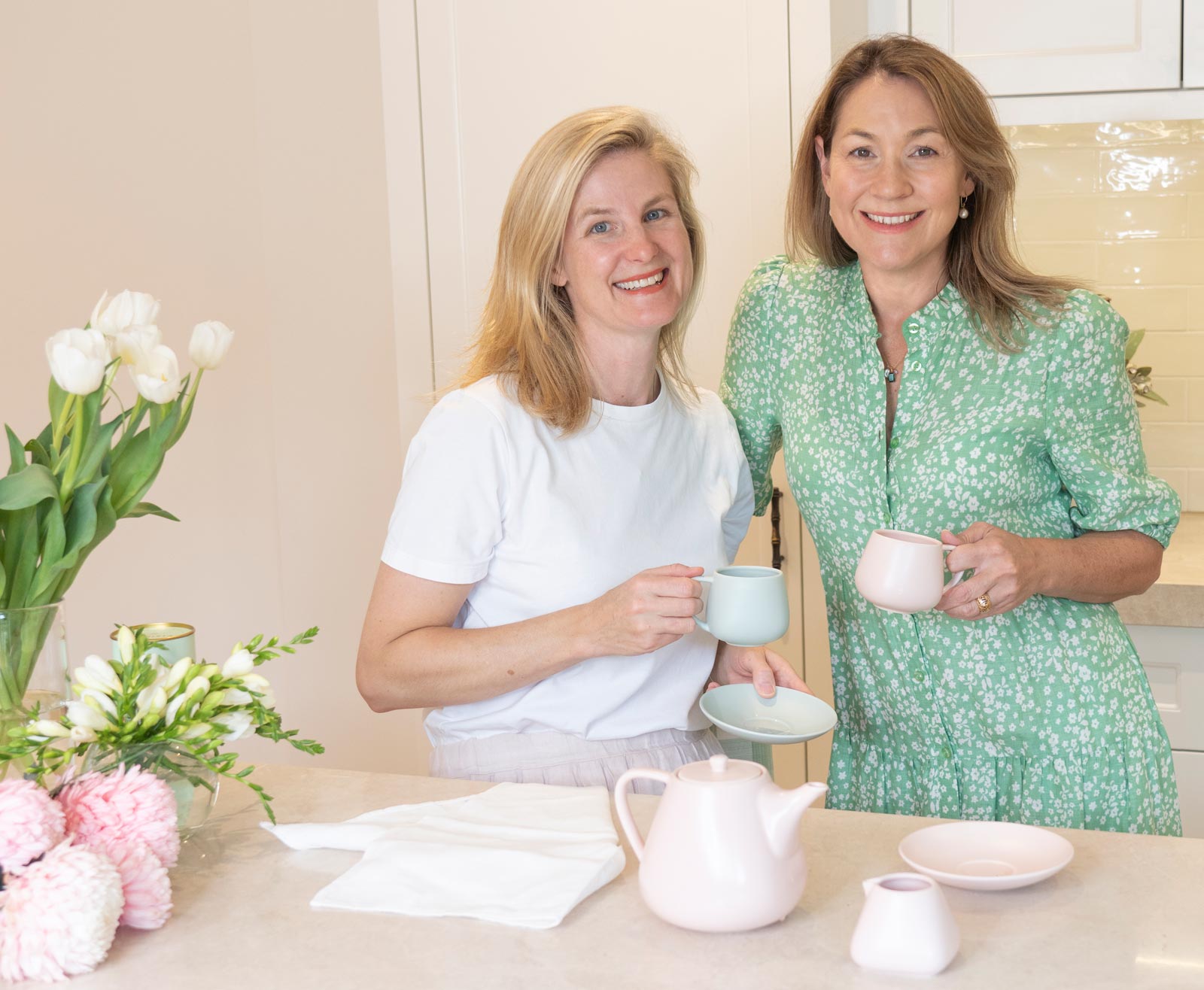 Two women smiling in a bright kitchen setting, holding pastel-colored teacups. On the table in front of them are matching pastel teapots, cups, saucers, and folded white napkins, with flowers in the background.