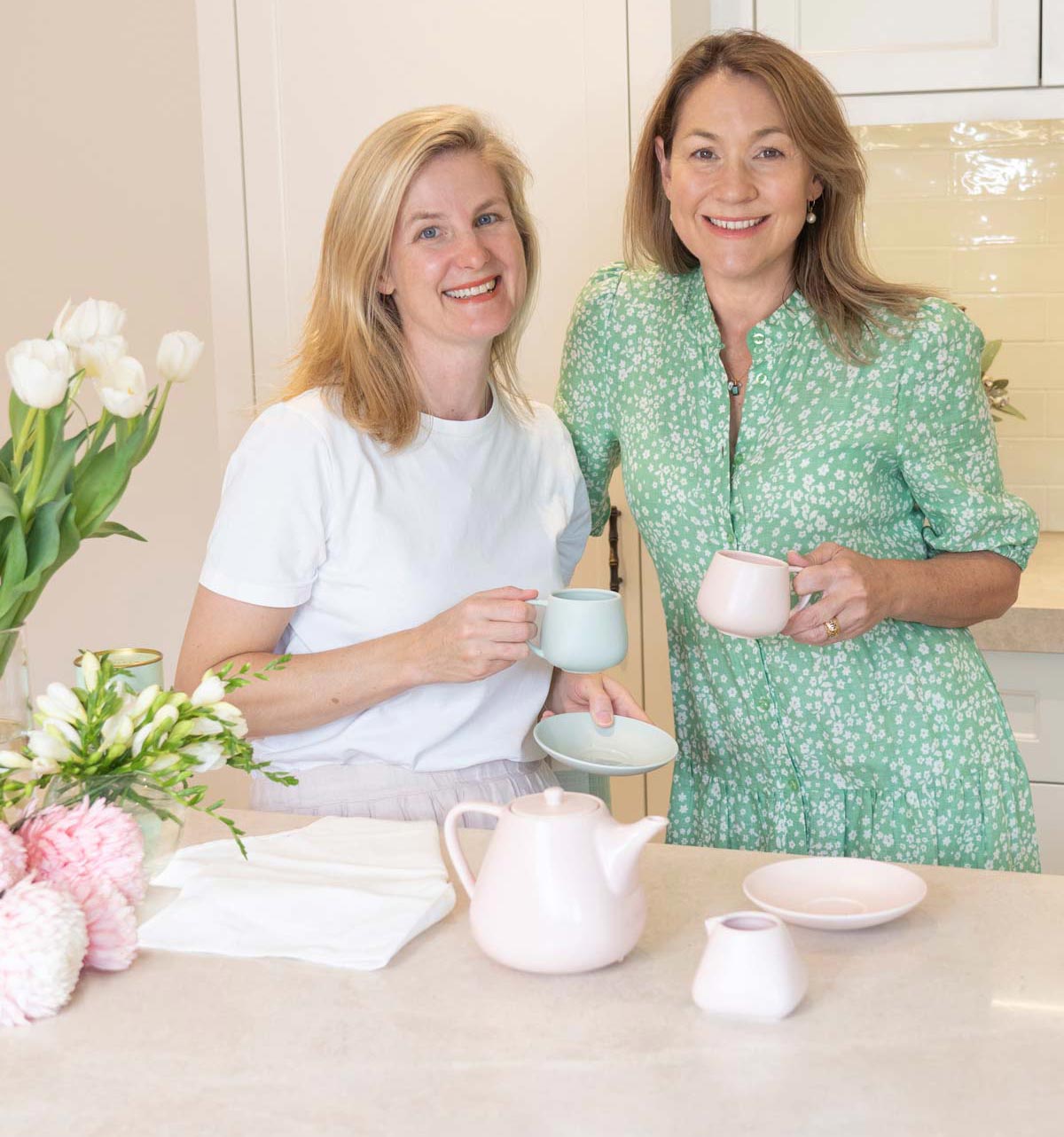 Two women sitting at a table with tea cups and a teapot, surrounded by flowers.
