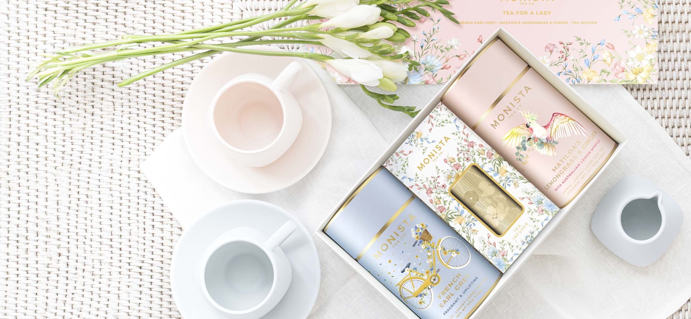 A styled flat-lay of a tea gift set featuring three pastel-colored canisters of tea with gold foil accents, displayed next to white ceramic teacups and a floral box of flowers on a white textured background.