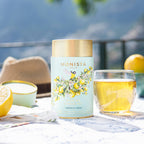 Sicilian Citrus Tea canister on an outdoor table with lemon halves, a glass of brewed tea, and a sun hat