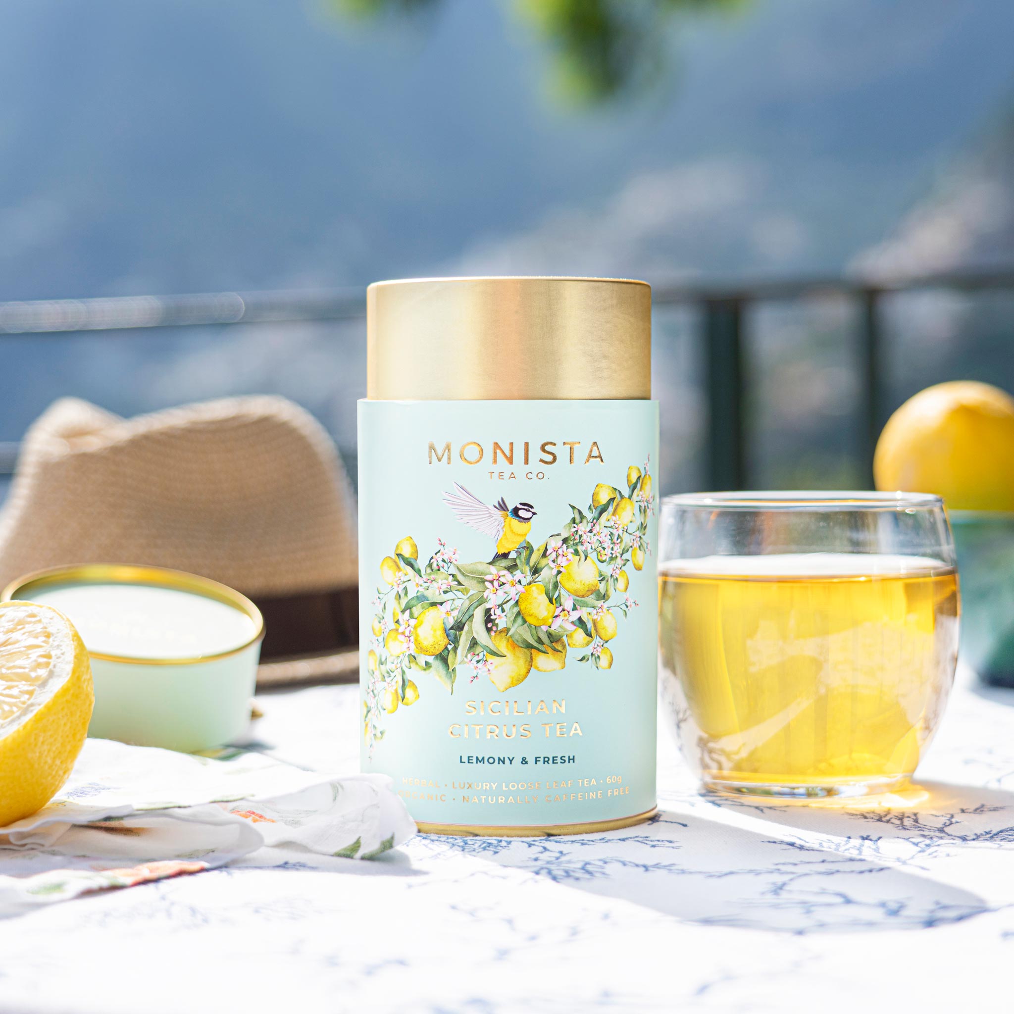 Sicilian Citrus Tea canister on an outdoor table with lemon halves, a glass of brewed tea, and a sun hat