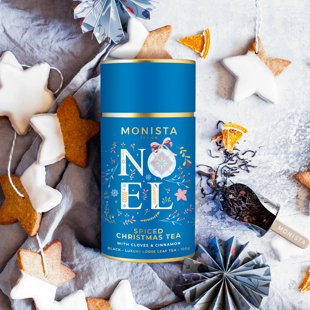 Monista Noel spiced Christmas tea canister surrounded by festive decorations on a textured surface.