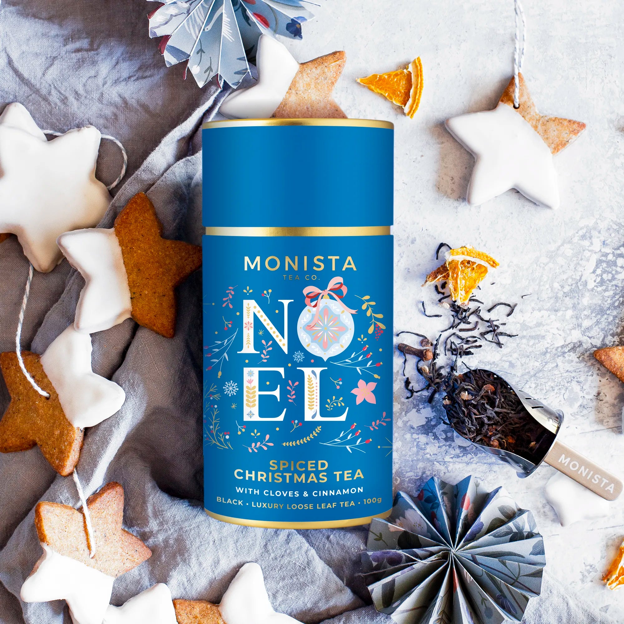 Monista Noel spiced Christmas tea canister surrounded by festive decorations on a textured surface.