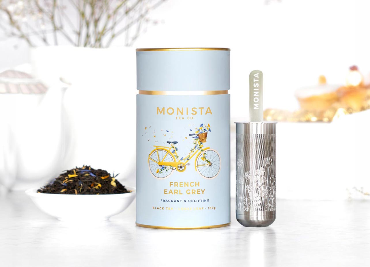 Monista French Earl Grey tea set featuring a light blue cylindrical tin with a yellow bicycle illustration, a silver metal infuser, and a small bowl of loose tea leaves on a white surface.