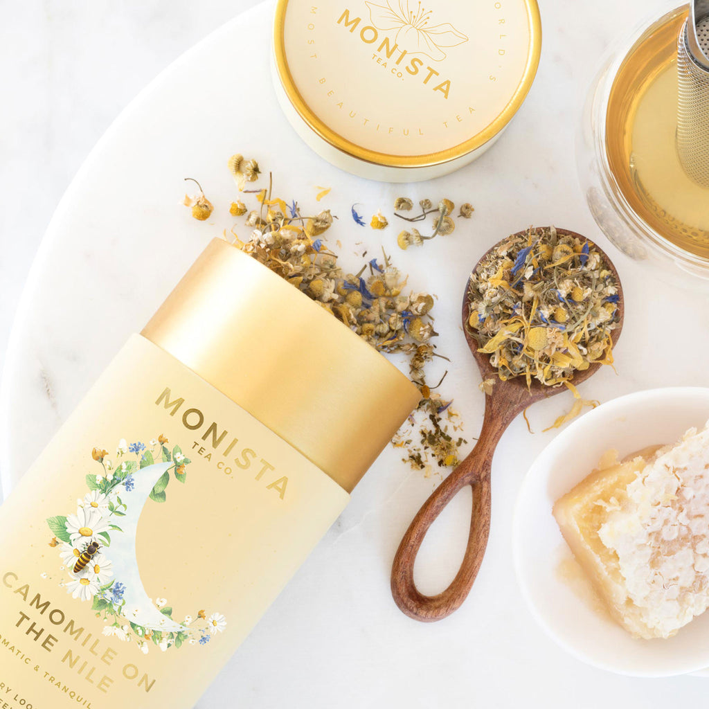 Camomile On The Nile tea canister with dried chamomile flowers, a gold spoon, and honeycomb on a marble surface