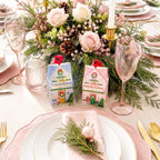 Two small Christmas-themed gift boxes with illustrated holiday house designs are placed on a styled table setting with plates, pink napkins, champagne glasses, and a floral centerpiece.