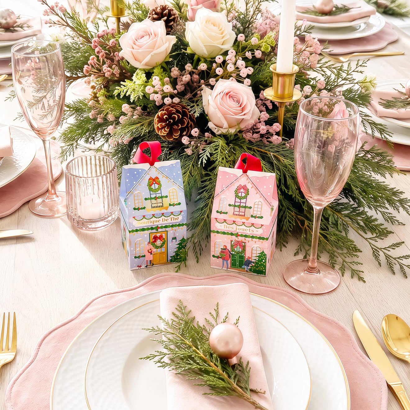 Two small Christmas-themed gift boxes with illustrated holiday house designs are placed on a styled table setting with plates, pink napkins, champagne glasses, and a floral centerpiece.