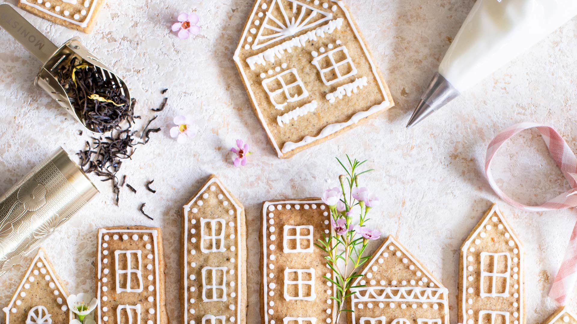 Decorated gingerbread house cookies arranged on a light surface with icing, a piping bag, loose tea leaves, and small pink flowers.