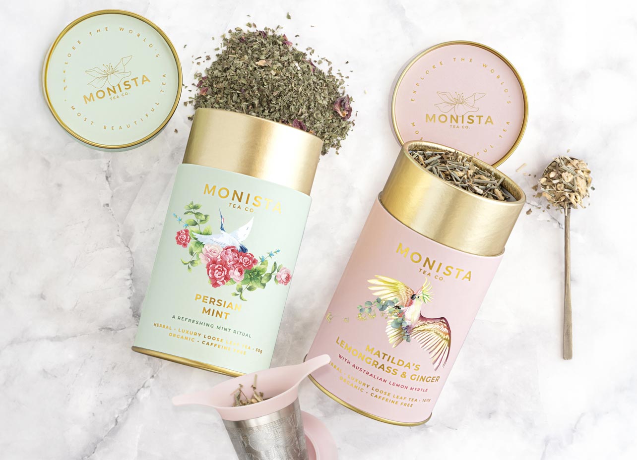 Two Monista tea tins with gold lids and gold lettering, displayed on a white marble surface with loose tea and a spoonful beside them. One tin is pale mint green labeled Persian Mint, and the other is pale pink labeled Matloa's Lemonbalm & Ginger.