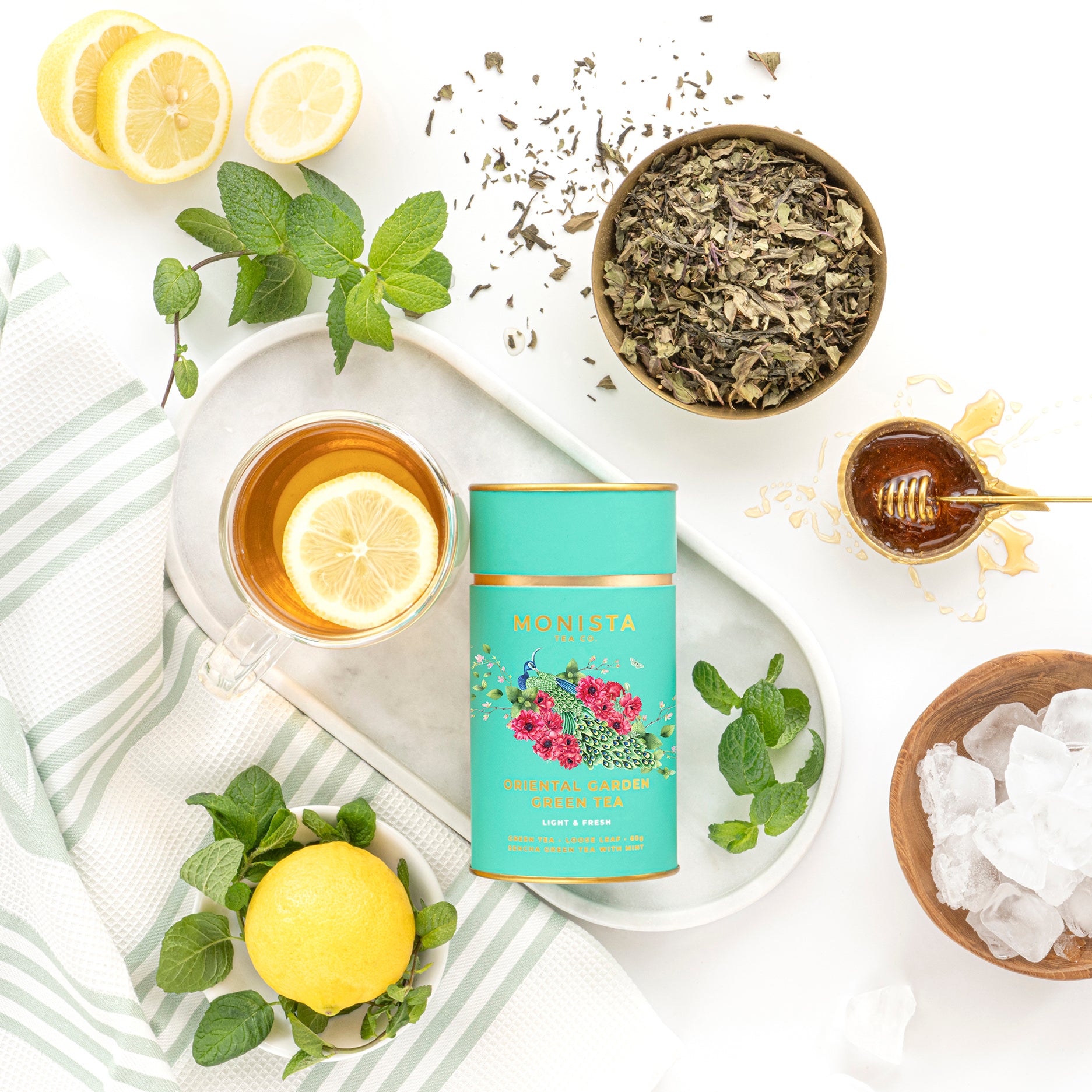 Oriental Garden Green Tea canister with lemon slices, mint leaves, loose tea, honey, and ice on a marble surface