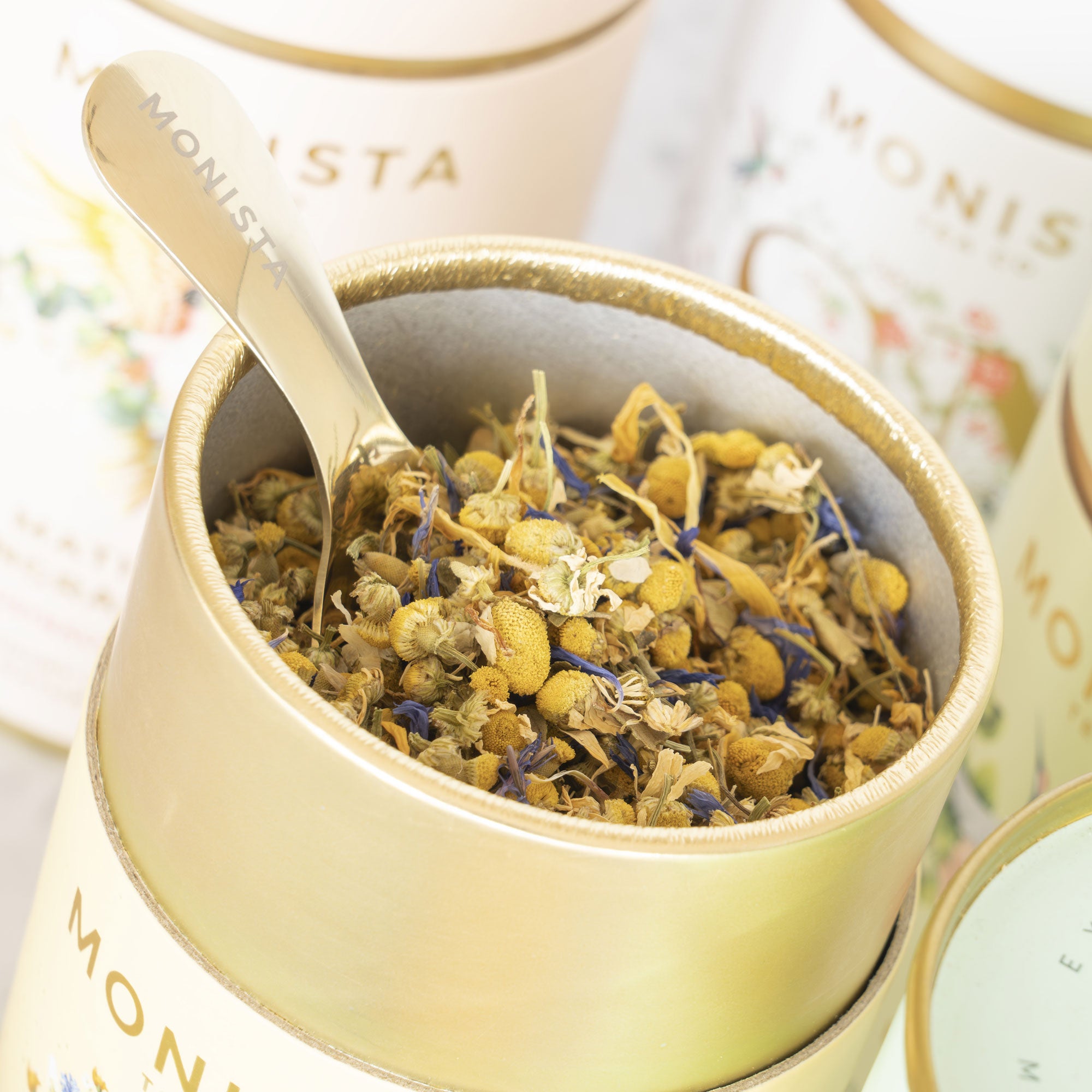 Gold spoon scooping dried chamomile flowers from a gold tea canister
